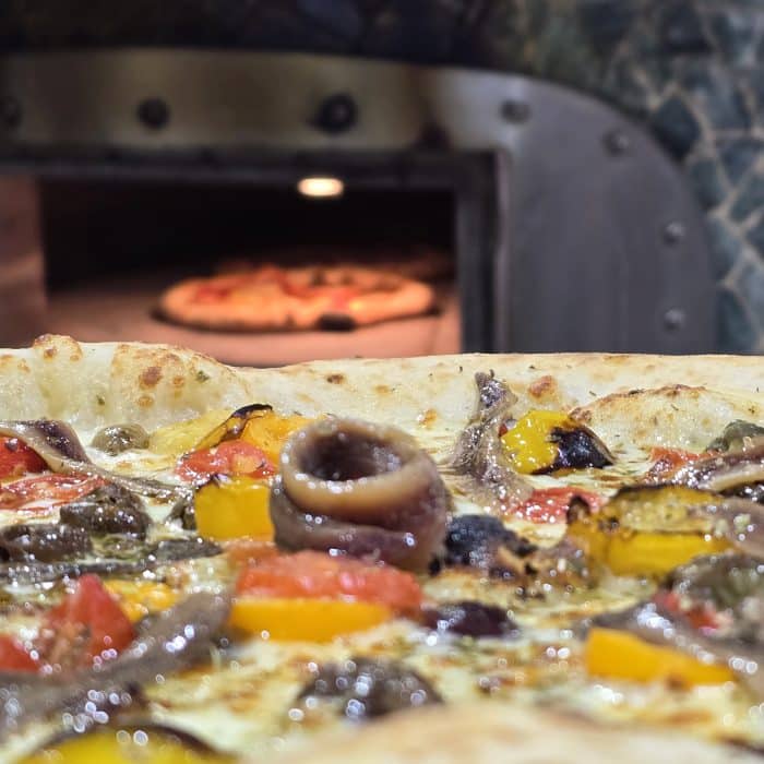 Une pizza aux câpres vue de près avec un four à pizza en fond